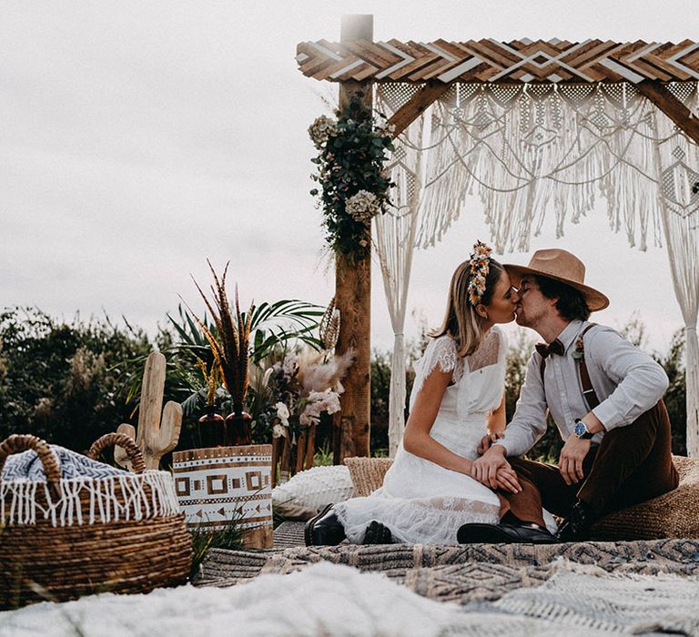 Bride in a lace ruffle wedding dress sitting on cushions with the groom in a striped shirt and brown hat for their outdoor ceremony 