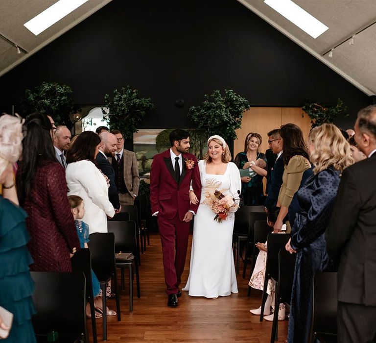 The groom in a burgundy suit with black lapels walks with the bride down the aisle together with the bride in a long sleeve wedding dress 