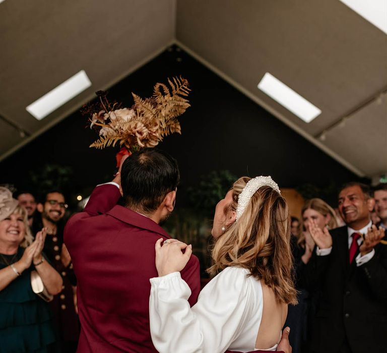 The groom in a burgundy suit and bride wearing pearl accessories turn and face the guests as a married couple 