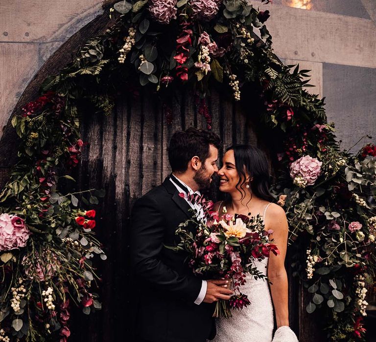 Bride in beaded sparkly wedding dress smiling with the groom under the pink wedding flower arch arrangement 