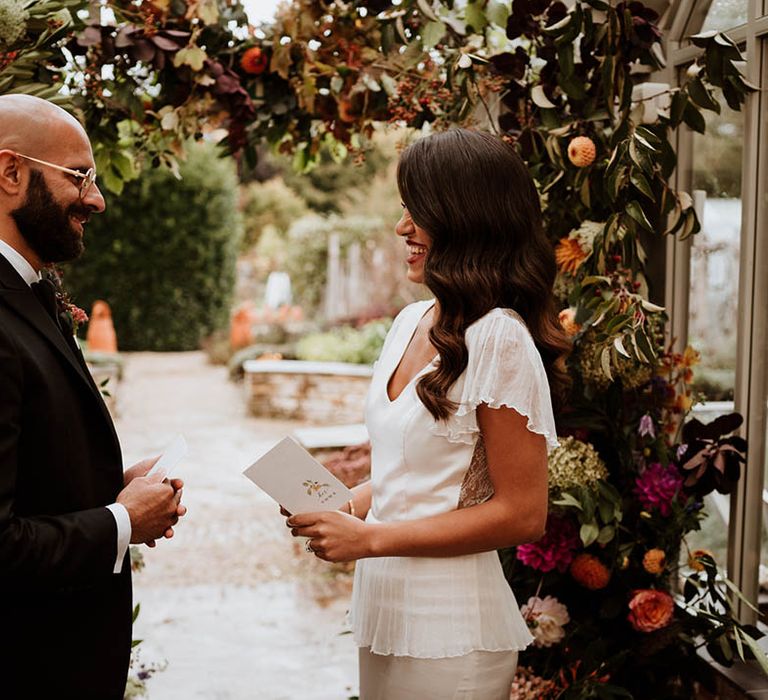 Bride and groom reading their vows at La Manoir Aux Quat Saisons standing by large floral arch with white and pink carnations, garden roses, red strawflower, eucalyptus, baby’s-breath and various dried flowers 