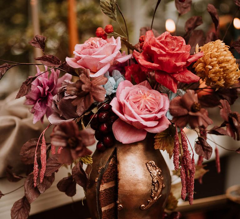 single tier iced wedding cake with intricate leaf details in the style of an aged golden vase filled with handmade wafer paper florals, pink roses, red roses and yellow carnations 