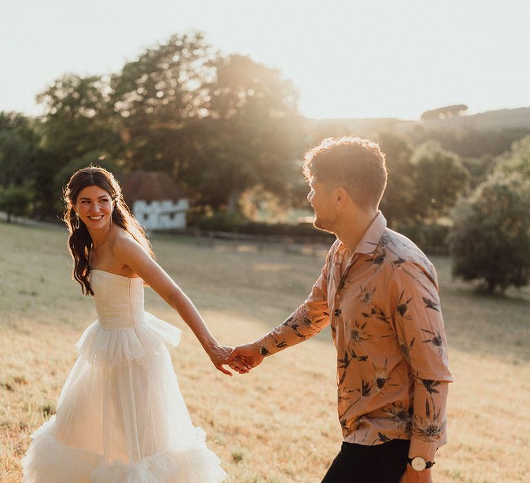 Groom in pink summer shirt with thistle design holding hands with the bride in a strapless ruffle wedding dress during golden hour 