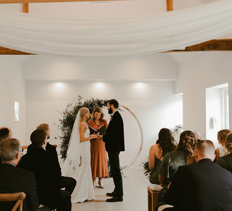 Bride and groom stand holding hands at the altar at Southend Barns with a floral moongate 