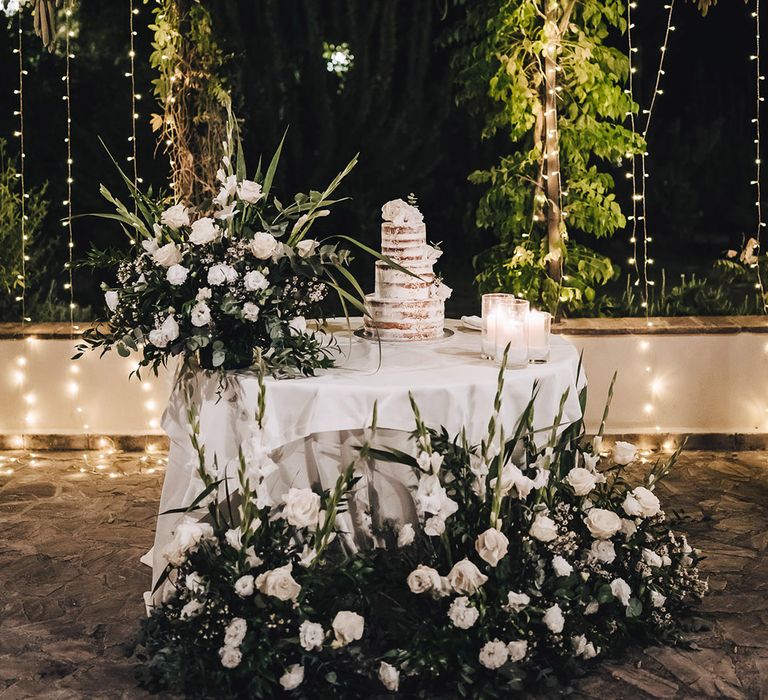 Large white floral arrangements with green foliage beside round table complete with naked wedding cake