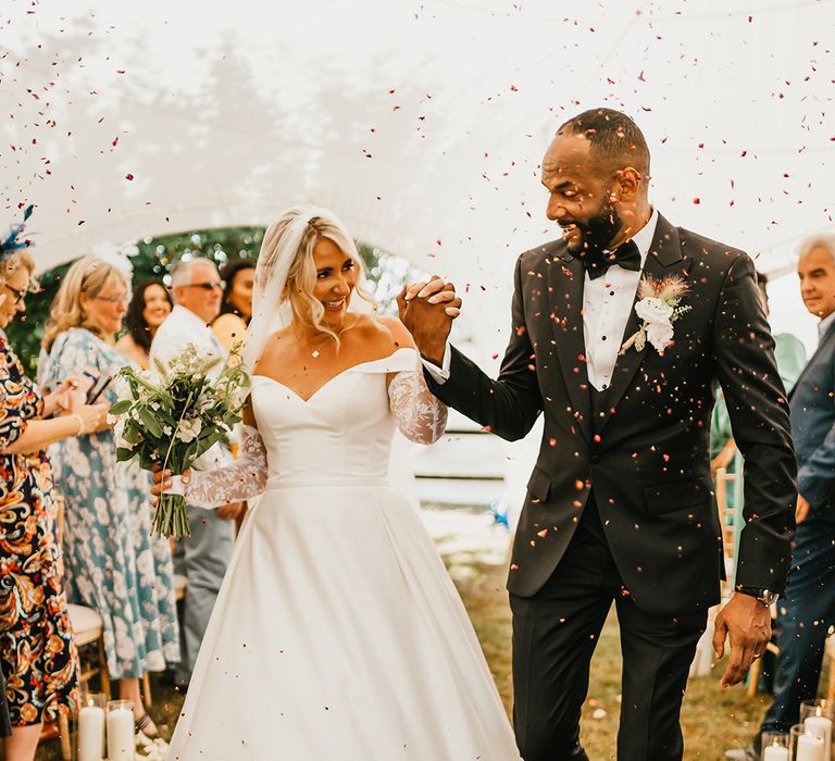 Bride in princess wedding dress and detachable lace sleeves walks through colourful confetti exit with her groom in black-tie