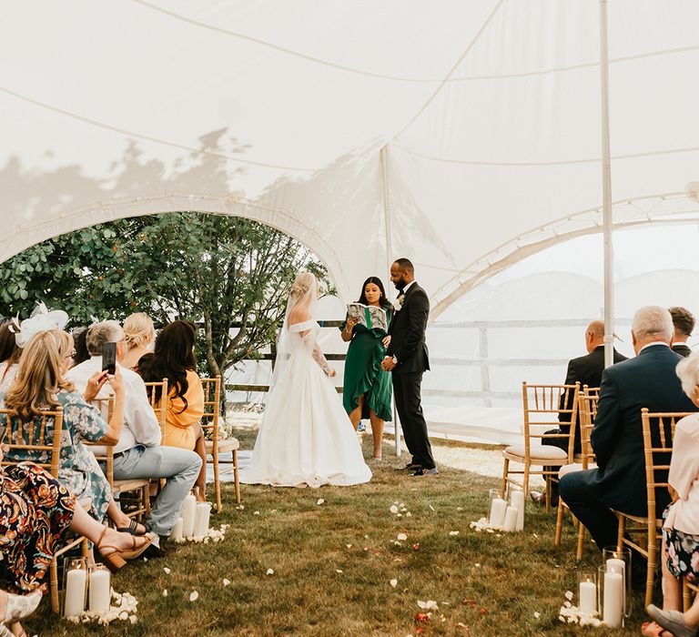 Bride in floor-length veil stands opposite her groom in black-tie during tent ceremony for back-garden wedding