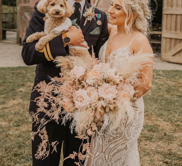 The bride and groom smile together holding their pet dog, Nala on their wedding day wearing a cute collared white dog outfit 