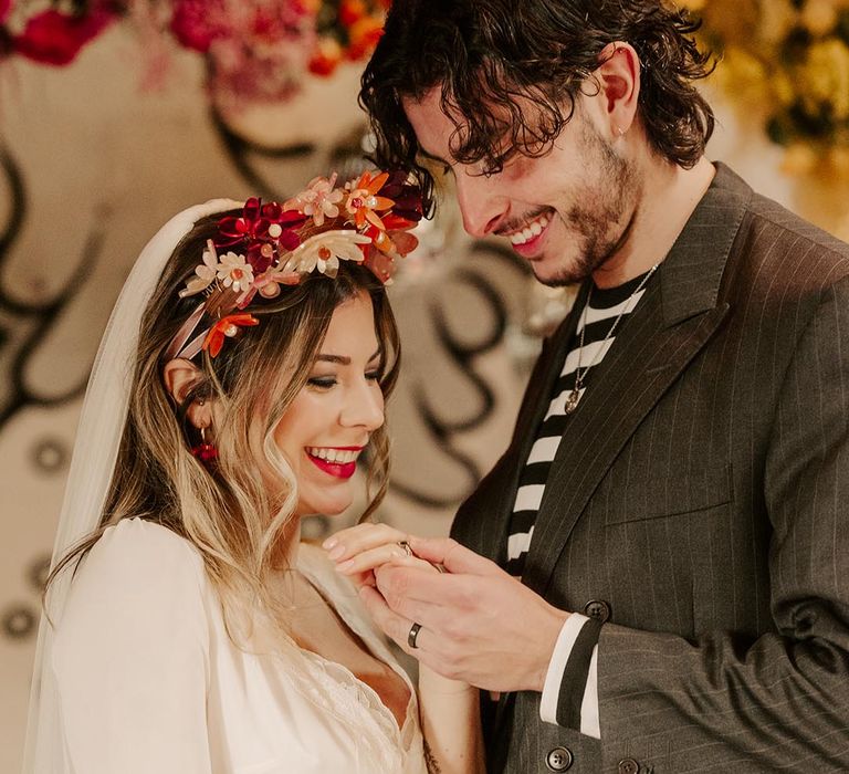 Bride in three quarter length sheer sleeve wedding dress with button and cinched waist details and pink bridal flower crown standing with groom in dark grey pinstripe blazer and black and white long sleeve t shirt 