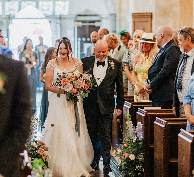 Father of the bride in black tie walks down the aisle with the bride in a flower wedding dress walking down the aisle to the groom at the end 