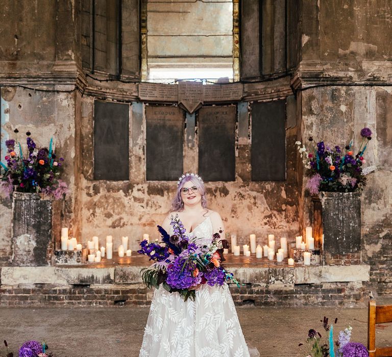 Bride in leaf appliqué sheer Evie Young bridal gown and cathedral length veil standing in Asylum Chapel London holding bright purple bridal bouquet with purple hydrangeas, pink garden roses, purple orchids, lilac carnations, dried flowers and peacock blue feathers surrounded by pillar candles and purple, blue, pink and white flower arrangements with dried flowers and bunny ears