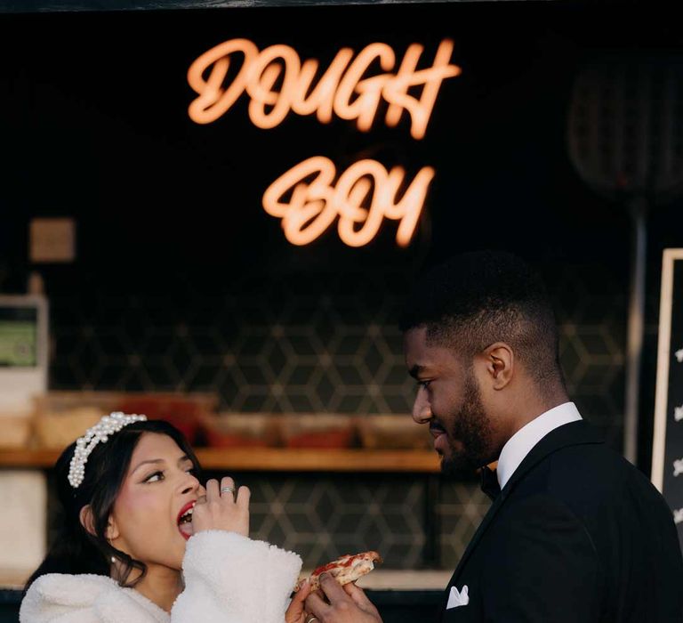 Bride in white fluffy coat and large pearl headband eating pizza with groom in classic black tuxedo and white pocket square 