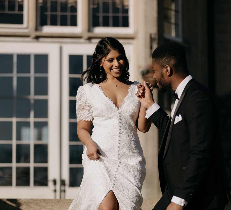 Bride in v neck short sleeve wedding dress with front slit and button detailing and pearl detailed wedding heels walking down the steps with groom in classic black tuxedo with white pocket square 