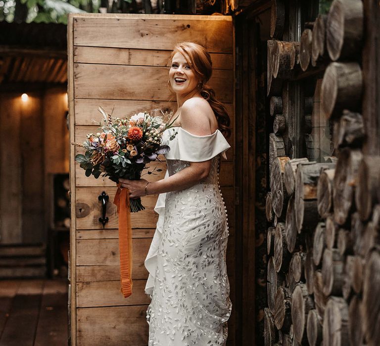 Bride off-the-shoulder floral embellished Love Spell wedding dress and carries Autumnal floral bouquet 