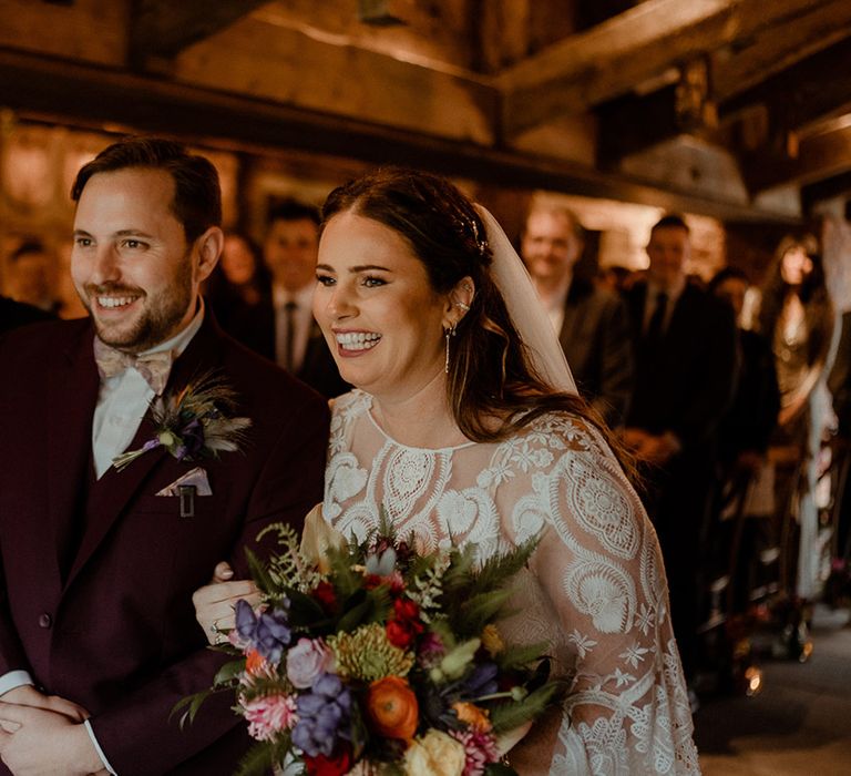 Bride in boho lace wedding dress stands at the altar with the groom in a burgundy suit and paisley bow tie with a dried flower buttonhole with a peacock feather 