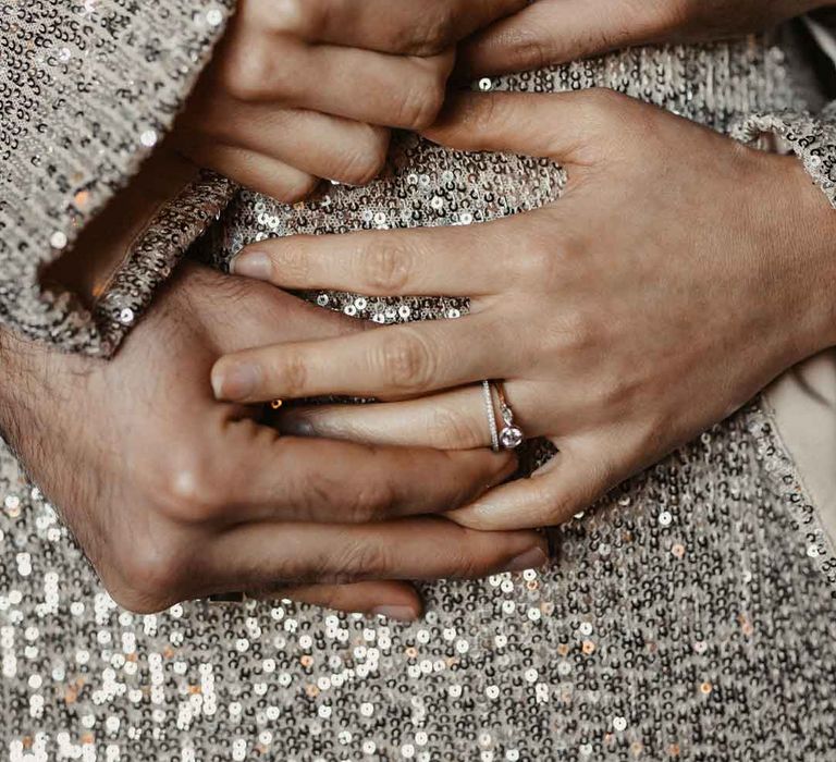 Modern luxe wedding rings holding hands against silver sparkly dress 