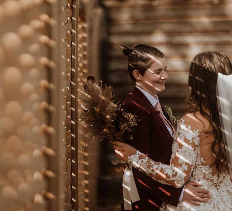 Couples wedding portraits - bride in maroon bridal suit with grey waistcoat, dusky pink tie and autumnal dried flower boutonniere embracing bride in long sleeve lace illusion sleeve wedding dress with overlay holding large autumnal boho dried flower bouquet 