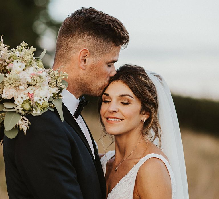 Groom in black tie kisses the bride on the forehead with the bride in a fitted beaded wedding dress with white flower wedding bouquet 