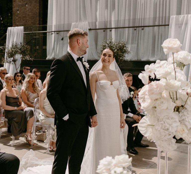 Bride in Alena Leena Bridal wedding dress standing with the groom in a black tuxedo for the outdoor city wedding 