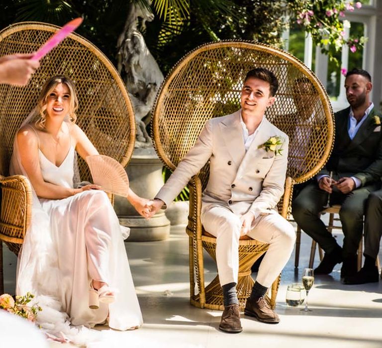 Bride in satin bridal two piece with groom in double breasted cream suits sitting on peacock chairs 