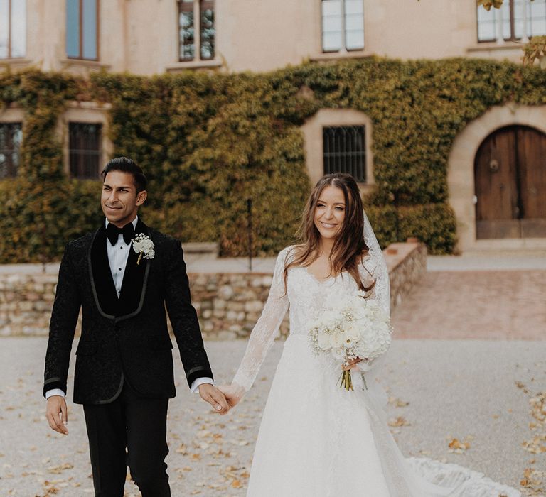 Bride in lace long sleeve Pronovias wedding dress and groom in classic black tuxedo with white flower boutonniere walking around venue 