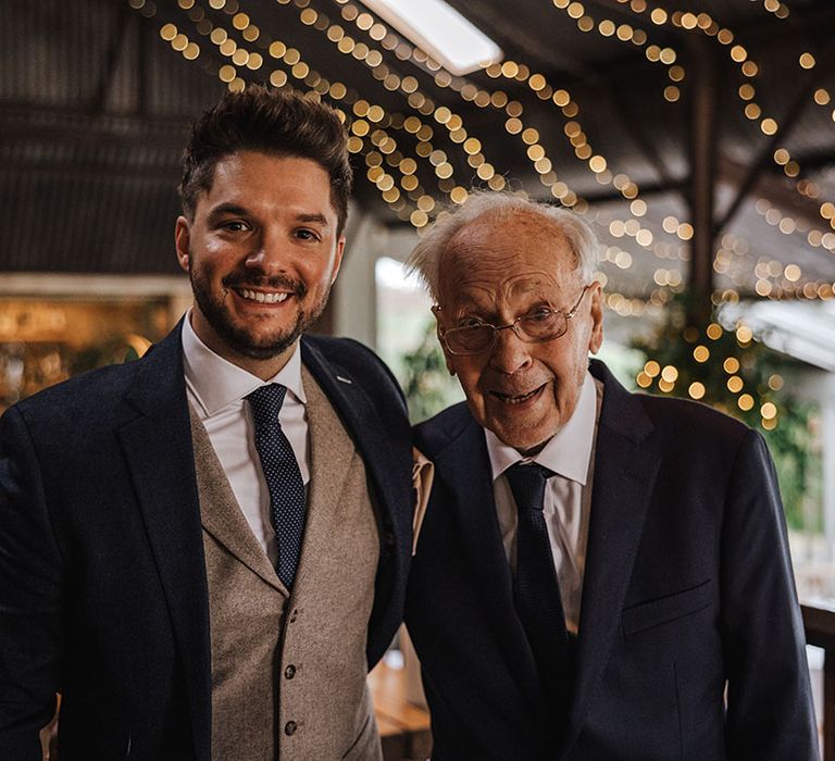 Groom in navy suit and tie with brown waistcoat smiling with grandad in navy suit 