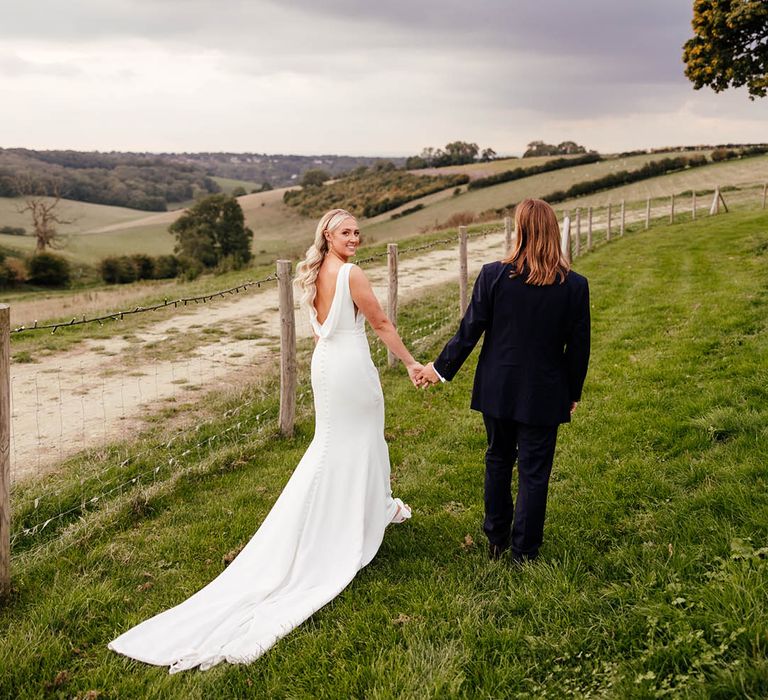 Bride with Hollywood curled blonde hair in draped back wedding dress walking hand in hand with the groom in blue suit 