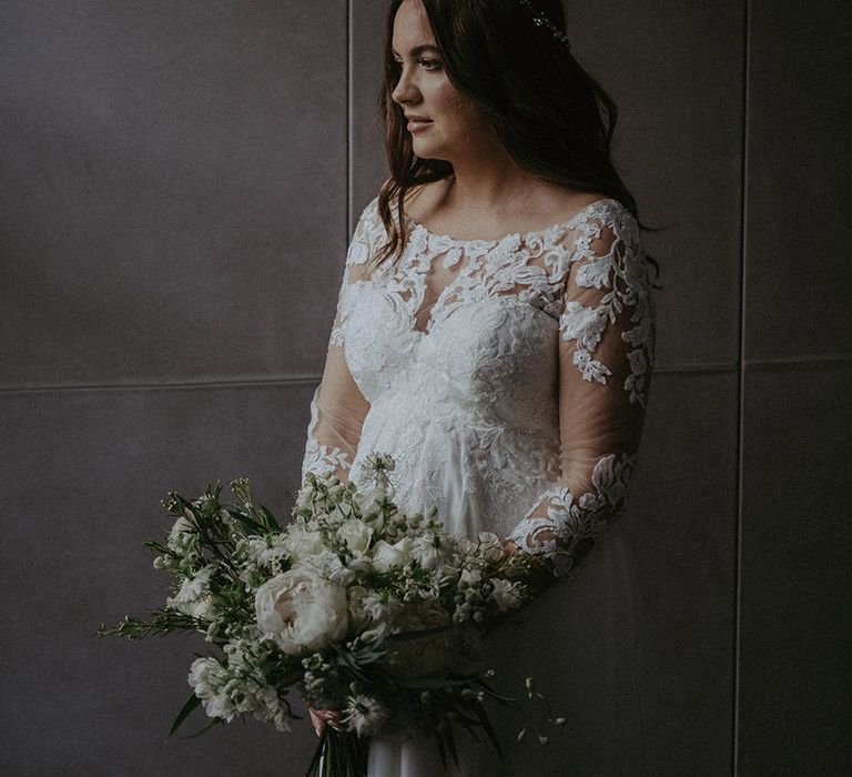 Bride in Pronovias lace long sleeve maternity wedding dress holding an all white wedding bouquet with brown hair in a curled style with sparkly hair accessory 