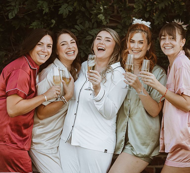 Bridesmaids in satin mismatched pyjamas with the bride in white pyjamas drinking champagne as they get ready for the classic wedding 