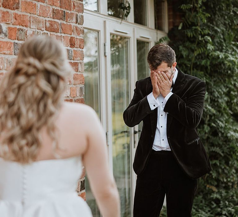 Groom in black velvet tuxedo puts his head in his hands crying as he gets the first look at the bride 