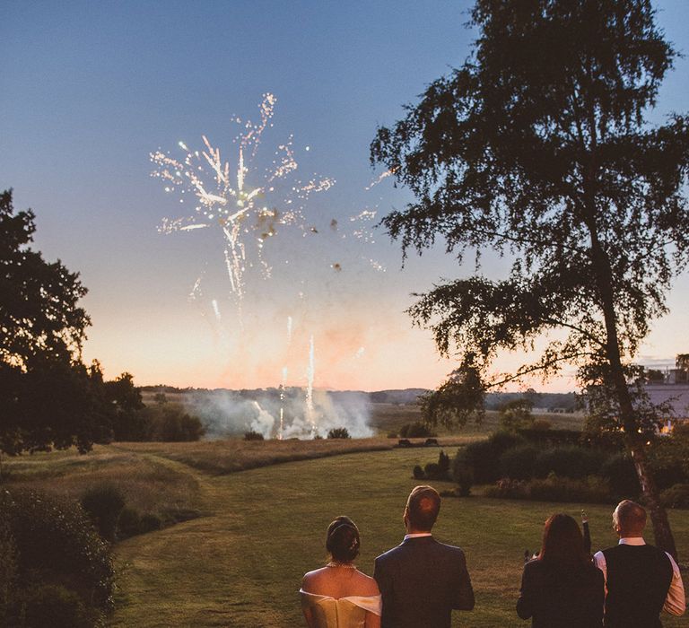 Bride in off the shoulder wedding dress with groom in navy blue suit looking at the firework display 