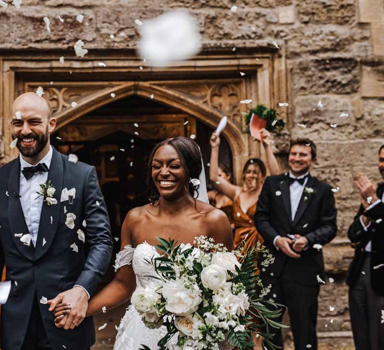 Bride in sweetheart neckline lace wedding dress with detachable off the shoulder sleeves leaving the church with groom in classic black three piece grooms suit with bow tie and white rose and foliage boutonniere doing dried petal confetti exit 