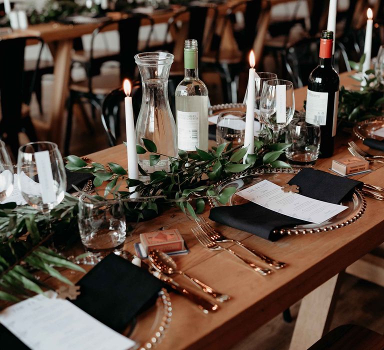 Wedding tablescape with green foliage, white tapered candles, black napkins and wine wedding drinks