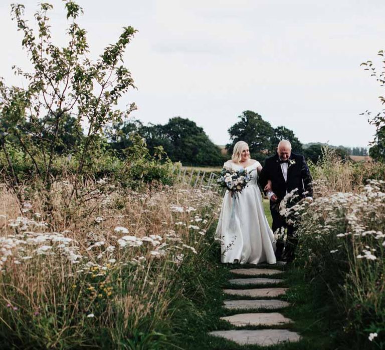 Bride in satin off the shoulder wedding dress holding eucalyptus, foliage, garden rose, gardenia, bluebell and dried flower bridal bouquet tied with dusky blue ribbon walking through the vineyard wedding venue with father of the groom