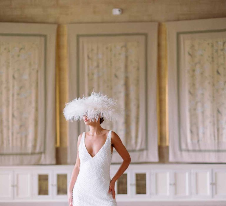 Bride in low v-neck slip bridal dress with large artistic feather bridal hat accessory at Settrington Orangery