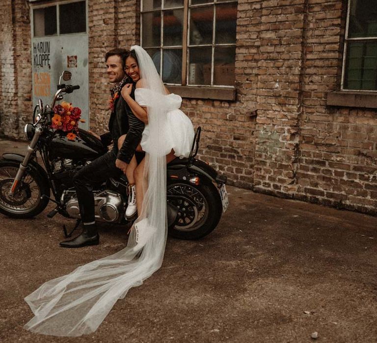 Bride in off shoulder tulle short wedding dress with church length veil and white platform Dr Martens wedding shoes on the back of wedding motorbike with groom in wet look black grooms suit with leopard print shirt and pink and orange boutonniere