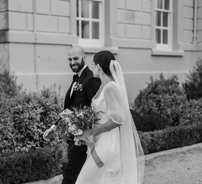 Bride in short sheer puff sleeve wedding dress with square neck and wrist loop with sheer pearl embellished bridal veil holding dried wildflower bouquet walking with groom in classic black tuxedo with bowtie and wildflower boutonniere at Buxted Park