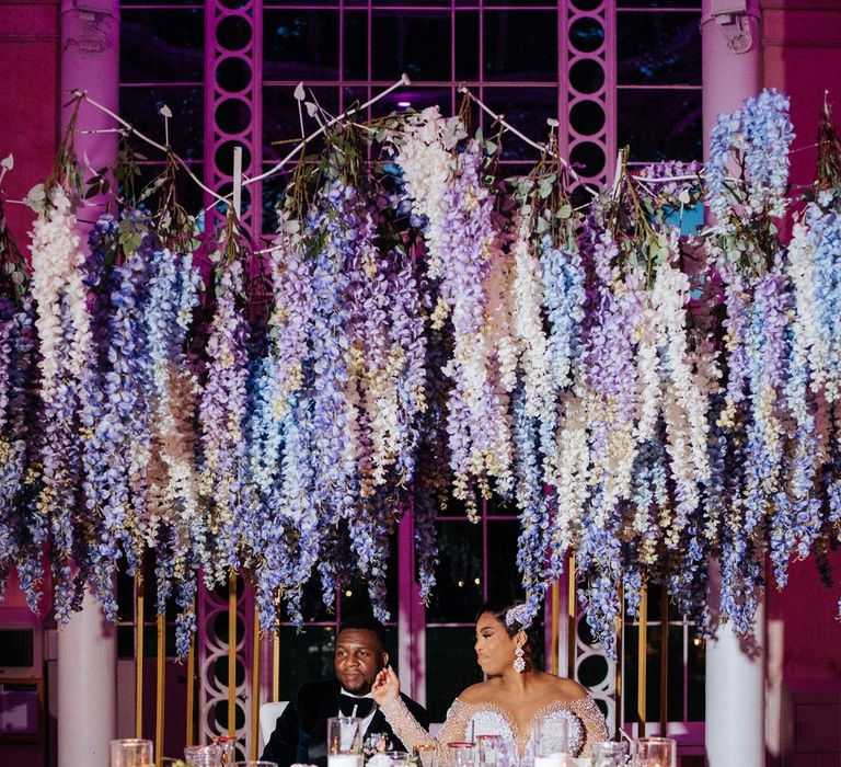 Bride in illusion v-neck sheer off shoulder long sleeve pearl embellished wedding gown sitting with groom in velvet midnight blue double breasted groom’s blazer, velvet bowtie and peach garden rose boutonniere at wedding top table with suspended pastel flower decor, hurricane vases and pillar candles, gold flower vases, pastel flower centrepieces and pastel and foliage flower arrangements 