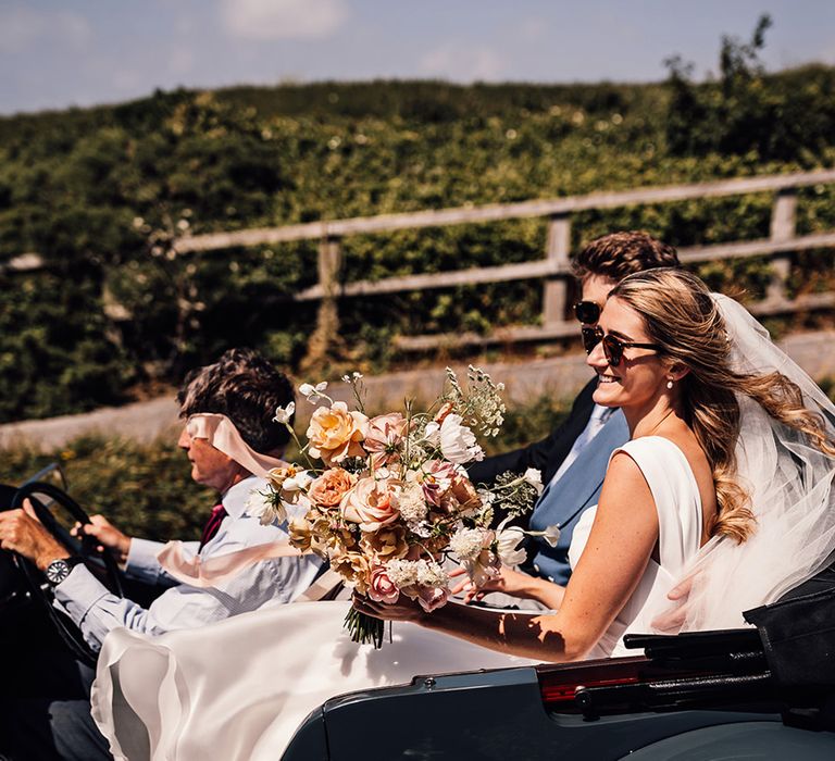 bride riding in a vintage convertible wedding car holding her blush bouquet at Sennen Cove wedding 