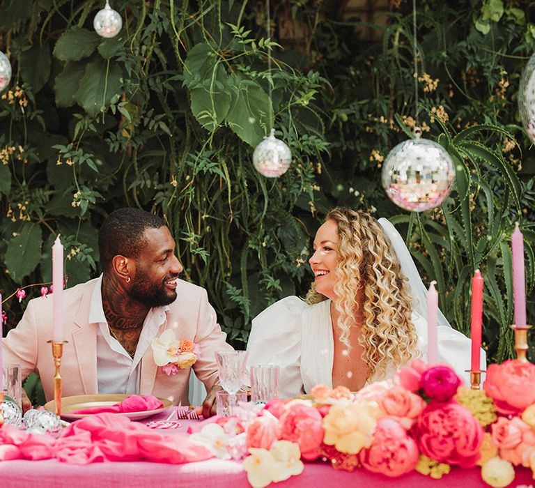 Bride and groom laughing at sweetheart table with disco ball decorations and pink styling 