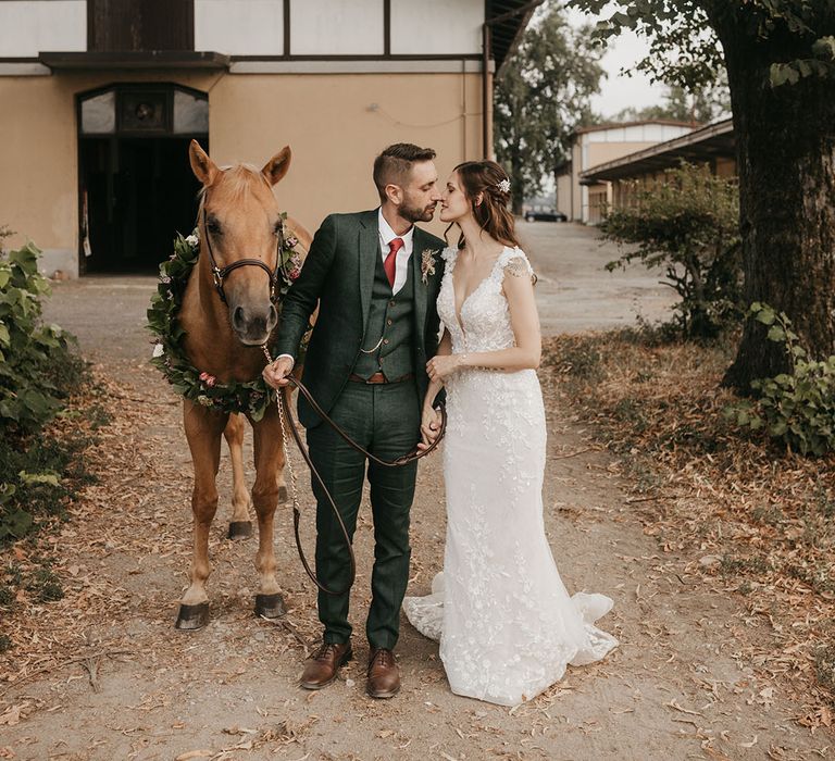 bride in an appliqué wedding dress kissing her groom in a dark green suit at riding school wedding with their pet horse in a flower collar next to them. 