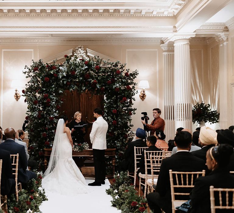 Classic and traditional Hedsor House wedding with red rose flower arch with the bride and groom in black tie 