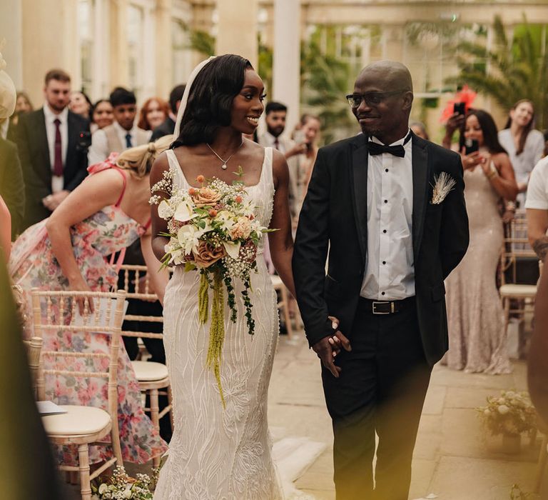 Father of the bride in a black tuxedo walks the bride in a beaded wedding dress down the aisle 