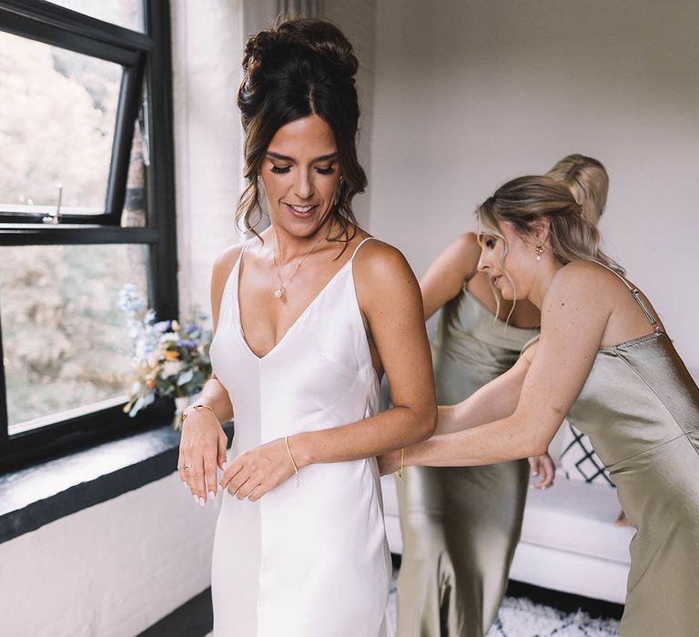 Bridesmaid in sage green satin bridesmaid dress helping bride into her satin wedding gown 