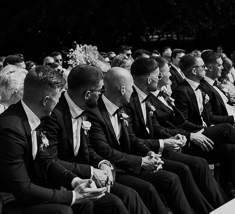 Groomsmen in black tuxedos sitting together at the ceremony 