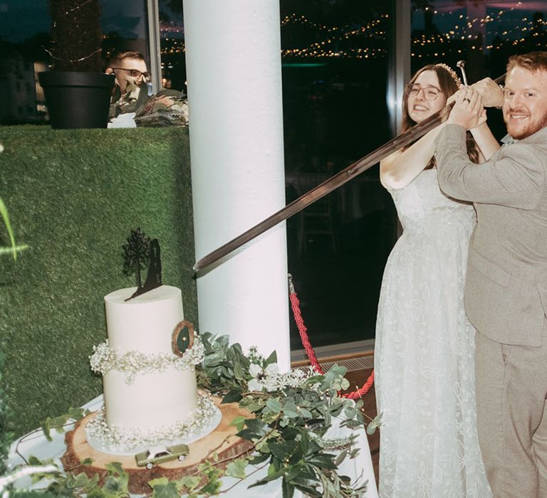 The bride and groom cut their two tier white frosted wedding cake with a large sword 