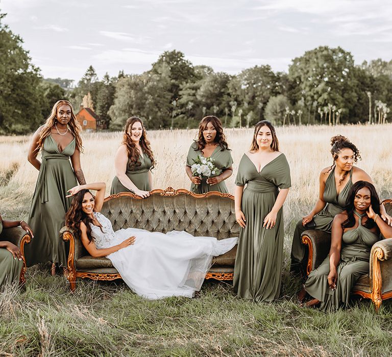 Bridal party posing on green lounges outdoors for group photo with green bridesmaid dresses and white rose bouquet