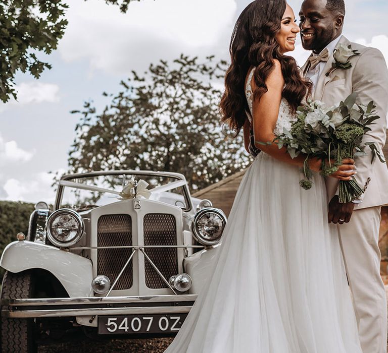 Bride in traditional princess wedding dress embracing groom in cream three piece suit in front of their vintage wedding car 