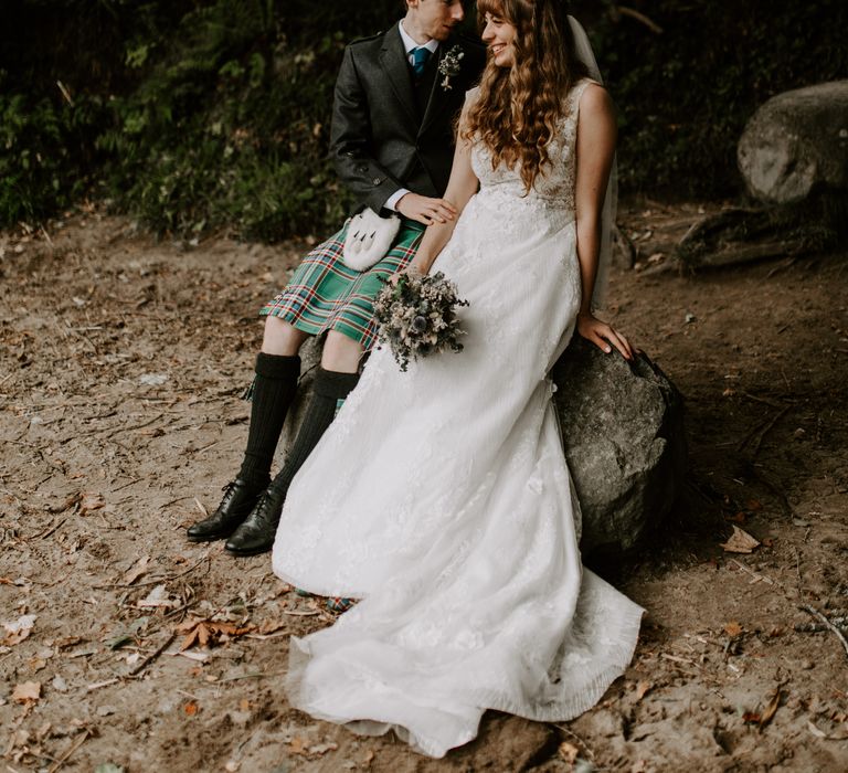 Bride and groom sit on rock in a forest in the Scottish Highlands with stunning dried flower bouquet