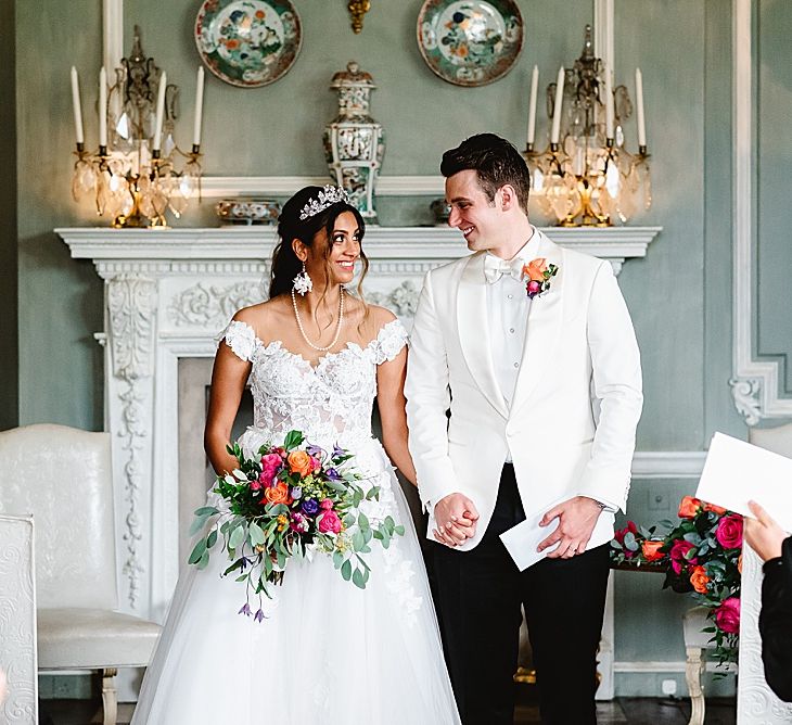 Bride in a Galia Lahav wedding dress and tiara and groom in a white tuxedo jacket and bow tie holding hands at the altar